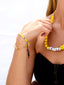 Close up of Venessa Arizaga SS26 Good Vibes Only Bracelet on wrist — adjustable black threadwork with multi-layer gold chains, glass pearls, and beaming smiley face charms; adjustable friendship bracelet design radiating handmade positivity, with Sugar Rush Necklace (Smile) made with leather cord, glass and ceramic charms with happy face draped over the collarbone.