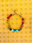 Venessa Arizaga FW26 Over The Rainbow Necklace - A colorful beaded statement necklace with rainbow gradient charms including heart, flower, smiley, clover, eyeball, mushroom, butterfly, bead, and ladybug in ceramic, wood, and glass on a yellow background.