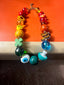 Venessa Arizaga FW26 Over The Rainbow Necklace - A colorful statement beaded necklace with rainbow gradient charms including heart, flower, smiley, clover, eyeball, mushroom, butterfly, bead, and ladybug in ceramic, wood, and glass, on a subway seat