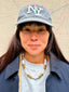 Model wearing a cap with 'NY' logo and Venessa Arizaga FW26 Talk of The Town Necklace - A statement gold beaded necklace with ceramic letter charms spelling "me", "you", "hi", "bye", "yes", "no", "omg" against a beige wall