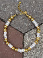 Lifestyle image of Venessa Arizaga FW26 Talk of The Town Necklace - A statement gold beaded necklace with ceramic letter charms spelling "me", "you", "hi", "bye", "yes", "no", "omg" on a textured surface