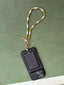 Lifestyle image of Venessa Arizaga FW26 Me Me Me Phone Leash - A statement gold beaded phone leash and bag charm with ceramic letter charms spelling "me". Black phone case with beaded lanyard on a green surface.
