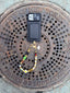 Lifestyle image of Venessa Arizaga FW26 Glitter Galaxy Phone Leash - A golden beaded phone strap/bag charm with ceramic and wooden charms in star, clover, bear, ladybug, butterfly, and flowers, perfect for making an everyday statement. Phone case with a colorful beaded necklace on a manhole cover with 'City of New York Parks & Recreation' text.