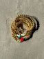 Lifestyle image of Venessa Arizaga FW26 Dreamwave Bracelet - Five rows of elastic gold beads and charms including a smiley face, ladybug, mushroom, alien, and star. Gold beaded bracelet with charms on a gray surface.