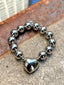 Lifestyle image of Venessa Arizaga FW26 Big Heart Bracelet (Silver) -  A silver beaded elastic statement bracelet with a large heart pendant. Silver beaded bracelet with a heart charm on a concrete surface.