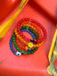 Lifestyle image of Venessa Arizaga FW26 Rainbow Reaction Bracelet  - A colorful five-row elastic bracelet with rainbow jade gradient beads and glass and ceramic charms including ladybug, smiley, mushroom, cactus, and eyeball, perfect for every day stacking. Colorful beaded bracelets on a red surface.