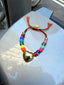 Venessa Arizaga FW26 Venus Heart Bracelet (Rainbow) - An everyday adjustable bracelet with rust threadwork, colorful beads, pave accents, and a gold heart pendant. Colorful beaded bracelet with a heart charm on a metal surface.