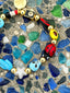 Venessa Arizaga FW26 Cosmic Roadtrip Bracelet - A golden beaded bracelet with classic icons - camera, smiley, butterfly, star, cactus, ladybug - perfect for everyday layering. Colorful beaded bracelet with various charms on a textured surface.