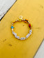 Venessa Arizaga FW26 High On Life Bracelet - A golden beaded bracelet with classic icons - smiley, flower, mushroom, hot air balloon, and butterfly - perfect for everyday layering. Bracelet with colorful beads and letter beads on a yellow and gray surface.