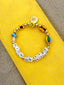Venessa Arizaga FW26 High On Life Bracelet - A golden beaded bracelet with classic icons - smiley, flower, mushroom, hot air balloon, and butterfly - perfect for everyday layering. Bracelet with colorful beads and letter beads on a yellow and gray surface.