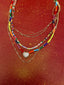 Lifestyle image of Venessa Arizaga FW26 Sweetheart Sparkle Necklace (Rainbow) - A statement layered necklace with rust threadwork, rainbow seed beads, gold plated chains, rhinestone chain, and a pave heart charm. Necklace with colorful beads on a red background