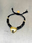 Lifestyle image of Venessa Arizaga FW26 Venus Heart Bracelet (Black) - An everyday adjustable bracelet with black threadwork, glass beads, pave accents, and a gold heart pendant. Black beaded bracelet with a gold heart charm on a gray surface.
