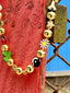 Lifestyle image of Venessa Arizaga FW26 Glitter Galaxy Necklace - A golden beaded necklace with classic icons - rainbow, smiley, alien, daisy, yin-yang, clover, star - perfect for everyday layering. Colorful beaded necklace with smiley face, evil eye, ladybug, four-leaf clover, and daisy pendants against a red wooden background.