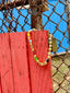 Lifestyle image of Venessa Arizaga FW26 Glitter Galaxy Necklace - A golden beaded necklace with classic icons - rainbow, smiley, alien, daisy, yin-yang, clover, star - perfect for everyday layering. Colorful beaded necklace with smiley face, evil eye, ladybug, four-leaf clover, and daisy pendants against a red wooden background.