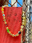 Lifestyle image of Venessa Arizaga FW26 Glitter Galaxy Necklace - A golden beaded necklace with classic icons - rainbow, smiley, alien, daisy, yin-yang, clover, star - perfect for everyday layering. Colorful beaded necklace with smiley face, evil eye, ladybug, four-leaf clover, and daisy pendants against a red wooden background.