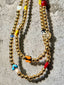 Lifestyle image of Venessa Arizaga FW26 Dreamwave Necklace - Two rows of gold beads and playful charms including a smiley face, ladybug, mushroom, alien, and star on a textured surface.