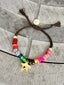 Lifestyle image of Venessa Arizaga FW26 Stardust Kiss Bracelet - A colorful adjustable bracelet with brown threadwork, colorful beads, pave accents, and a gold star pendant. Colorful beaded bracelet with star-shaped charm on a concrete surface