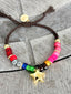 Lifestyle image of Venessa Arizaga FW26 Stardust Kiss Bracelet - A colorful adjustable bracelet with brown threadwork, colorful beads, pave accents, and a gold star pendant. Colorful beaded bracelet with star-shaped charm on a concrete surface
