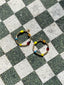 Lifestyle image of Venessa Arizaga FW26 Dreamwave Earrings - hoop earrings with gold beads and dangling butterfly, mushroom, and smiley charms on a checkered stone surface.
