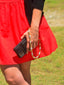 Model image of Venessa Arizaga FW26 Me Me Me Phone Leash - A statement gold beaded phone leash and bag charm with ceramic letter charms spelling "me". Person wearing a red dress and black sweater holding a phone with the phone leash.