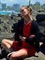 Close up model image of Venessa Arizaga FW26 Talk of The Town Necklace - A statement gold beaded necklace with ceramic letter charms spelling "me", "you", "hi", "bye", "yes", "no", "omg". Model sitting on rocks by the ocean wearing a red dress and black cardigan.
