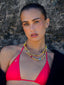 Model image of Venessa Arizaga FW26 Dreamwave Necklace - Two rows of gold beads and playful charms including a smiley face, ladybug, mushroom, alien, and star. Model wearing a red bikini top with a textured stone wall background.