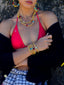 Model image of Venessa Arizaga FW26 Dreamwave Necklace - Two rows of gold beads and playful charms including a smiley face, ladybug, mushroom, alien, and star, the Dreamwave Bracelet - Five rows of elastic gold beads and charms including a smiley face, ladybug, mushroom, alien, and star, and Dreamwave Earrings - hoop earrings with gold beads and dangling butterfly, mushroom, and smiley charms . Model in colorful outfit standing against a textured wall.