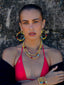Model image of Venessa Arizaga FW26 Dreamwave Necklace - Two rows of gold beads and playful charms including a smiley face, ladybug, mushroom, alien, and star, the Dreamwave Bracelet - Five rows of elastic gold beads and charms including a smiley face, ladybug, mushroom, alien, and star, and Dreamwave Earrings - hoop earrings with gold beads and dangling butterfly, mushroom, and smiley charms . Model in colorful outfit standing against a textured wall.