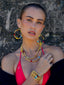 Model image of Venessa Arizaga FW26 Dreamwave Necklace - Two rows of gold beads and playful charms including a smiley face, ladybug, mushroom, alien, and star, the Dreamwave Bracelet - Five rows of elastic gold beads and charms including a smiley face, ladybug, mushroom, alien, and star, and Dreamwave Earrings - hoop earrings with gold beads and dangling butterfly, mushroom, and smiley charms . Model in colorful outfit standing against a textured wall.