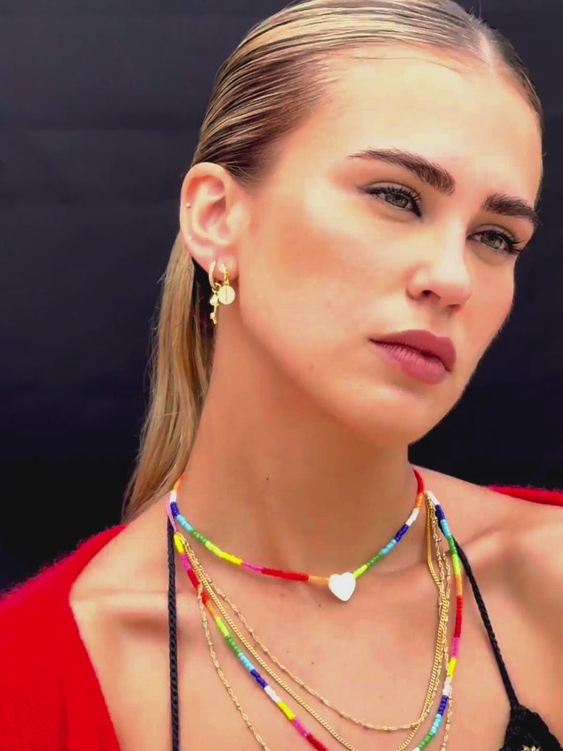Close up video of Venessa Arizaga SS26 Rainbow Aura Necklace layered at collarbone with the Rainbow Heartbeat Adjustable Necklace worn as a choker, styled with Peace Out Sparkle Earrings and Shimmer Huggie Hoops Pave earring with mushroom, perfect for a sun-drenched Paradisiac escape and effortless beach vibe.