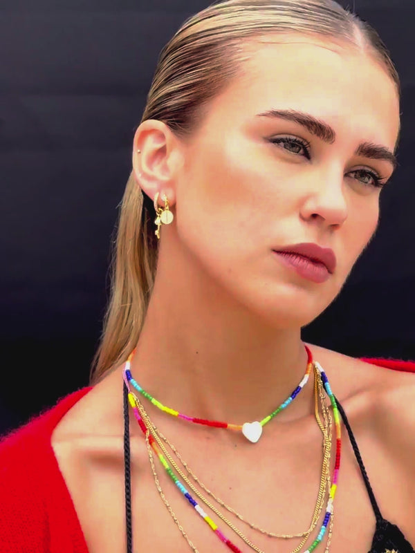 Close up video of Venessa Arizaga SS26 Rainbow Aura Necklace layered at collarbone with the Rainbow Heartbeat Adjustable Necklace worn as a choker, styled with Peace Out Sparkle Earrings and Shimmer Huggie Hoops Pave earring with mushroom, perfect for a sun-drenched Paradisiac escape and effortless beach vibe.
